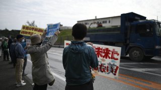 In this Feb. 25, 2019, photo, protesters with placards stand in front of the gate while construction vehicles enters the site during anti U.S. base protest outside of the U.S Base Camp Schwab gate in Nago, Okinawa prefecture, Japan. Based on a prefectural referendum result, more than 70% of voters in Okinawa reject U.S. base transfer in Henoko.