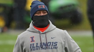 Illinois head football coach Lovie Smith, wearing a gray hooded sweatshirt, black gaiter mask and blue stocking cap, walks across the field before a game against Northwestern University