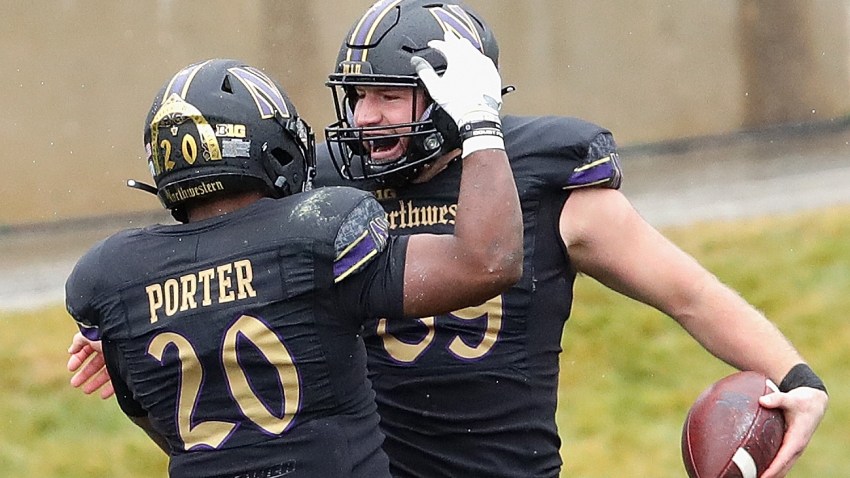 Two Northwestern University football players, wearing black uniforms with black helmets and gold trim throughout, celebrate a touchdown against Illinois in the annual rivalry game between the schools