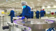 Workers in protective gear stand at machines putting vaccine into tiny vials