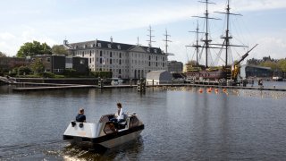 An electric boat steers close to a full-size replica of the 18th century three-mast trading ship Amsterdam at the National Maritime Museum, in Amsterdam, Thursday, May 20, 2021. Already steeped in maritime history, the city's more than 100 kilometers (60 miles) of waterways are to start hosting prototypes of futuristic boats — small, fully-autonomous electric vessels — to carry out tasks including transporting passengers and picking up garbage.