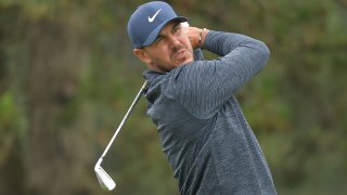 Golfer Brooks Koepka, clad in a blue hat and a blue windbreaker, finishes his swing with an iron during competition at the Travelers Championship