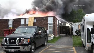 In the foreground, a black truck sits in front of a burning brick building, with flames shooting from the roof