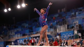 Simone Biles stands on the balance beam with her arms raised.