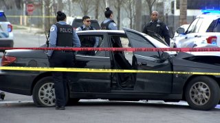 A black Toyota car is surrounded by red and yellow police tape, along with police officers, after its occupants were shot on April 11, 2022.