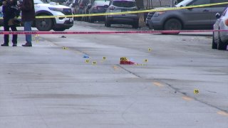 Yellow evidence markers lie on the scene of a shooting in Chicago in early January 2023. The scene is surrounded by yellow and red police tape, and Chicago police officers are seen in the background.