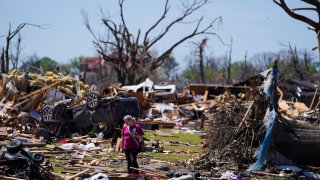 A woman walks near an uprooted tree, a flipped vehicle and debris from homes damaged by a tornado on March 27, 2023, in Rolling Fork, Miss.