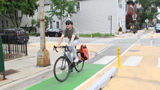 A man in a gray shirt and blue jeans rides a black and silver bicycle along a protected bike lane, with curbs on the sides and green paint in the middle, in Ukrainian Village.