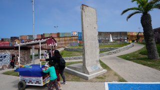 A family pushes a snack cart past a slab of the Berlin Wall, displayed near the border wall that separates the United States from Mexico, in Tijuana, Mexico, Friday, Aug. 25, 2023.