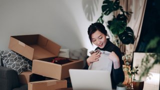 Young Asian woman unboxing new purchase clothings from cardboard box that received from her online shopping retail delivery at home. She is happy and excited to see the content from the box. Online shopping, trustworthy parcel delivery service