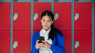 A medium close-up front view of a teenage girl who is sending text messages to a friend and checking her social media as she waits in the corridor of the high school she attends.