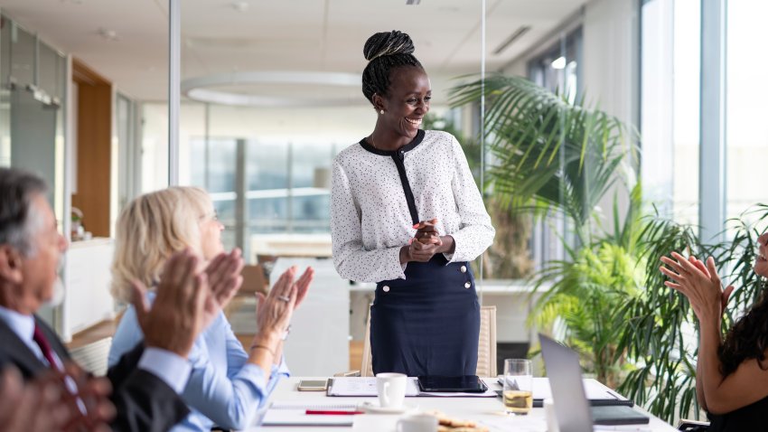 Front view of businesswoman in mid 30s smiling as colleagues applaud at conclusion of successful presentation.