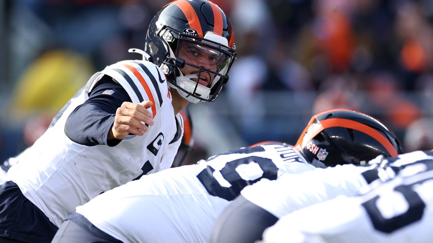 Bears quarterback Caleb Williams, wearing the team's blue-and-orange-striped helmet and white jerseys, points to a wide receiver on the side of the Bears' offensive line.