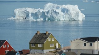 An iceberg floats in Disko Bay behind houses during unseasonably warm weather on July 30, 2019 in Ilulissat, Greenland. Climate change is having a profound effect in Greenland, where over the last several decades summers have become longer and the rate that glaciers and the Greenland ice cap are retreating has accelerated.