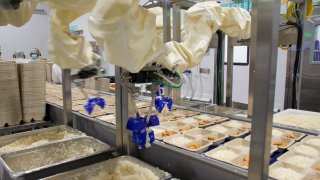 Chef Robotics builds long arms with multiple joints that are wrapped in a protective covering. The arms hang from metal racks that sit next to food-assembly lines and can scoop up different ingredients and spread them in prepackaged meal containers.