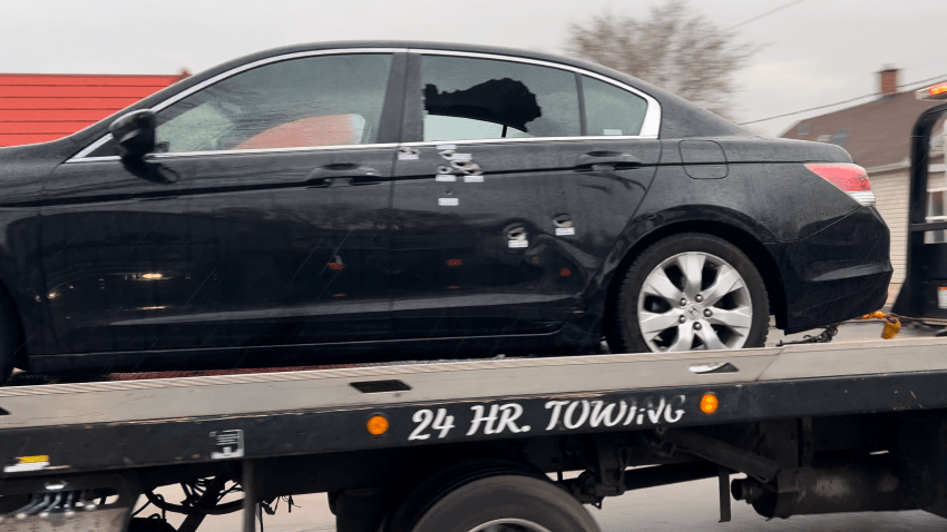 A black vehicle is shown on a tow truck. A window has been shot out and several bullet holes are visible in the rear door.