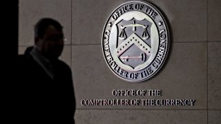 A pedestrian passes the seal of the Office of the Comptroller of the Currency displayed outside the organization’s headquarters in Washington, D.C., on March 20, 2019.