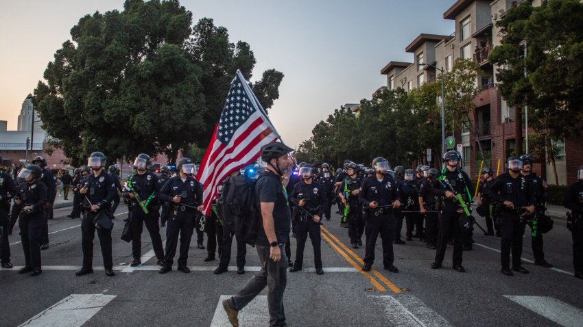 A man walks holding an American flag in front of the Los Angeles Metro Police while they try to disperse demontrators at the Little Tokyo neiborhood during protest in response to federal immigration operations in Los Angeles, on June 9, 2025. US President Donald Trump on June 9 ordered active-duty Marines into Los Angeles, vowing those protesting immigration arrests would be “hit harder” than ever. Protests in Los Angeles, home to a large Latino population, broke out on June 6, triggered by immigration raids that resulted in dozens of arrests of what authorities say are illegal migrants and gang members. (Photo by Apu GOMES / AFP) (Photo by APU GOMES/AFP via Getty Images)