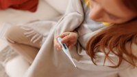 Sick young woman wrapped in a blanket, checking her temperature while sitting on a white sofa at home, seeking comfort and warmth during illness and recovery from a cold or flu