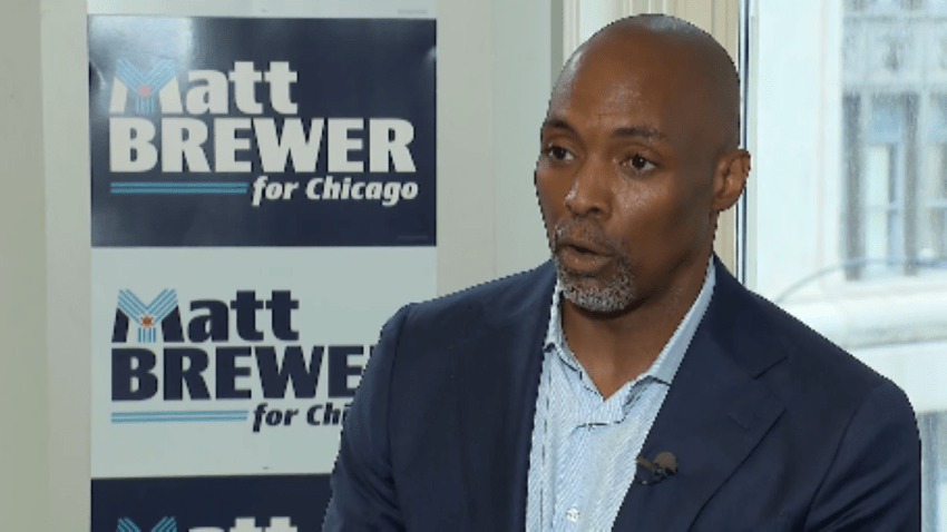 Chicago mayoral candidate Matt Brewer is pictured in front of multiple blue and white campaign signs. He's wearing a blue blazer and a light blue dress shirt.