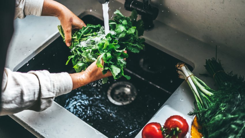 Hands rinsing fresh green leafy vegetables under flowing water in the sink, ensuring cleanliness for healthy cooking. Bright kitchen scene with tomatoes and herbs adding vibrancy to the setting.